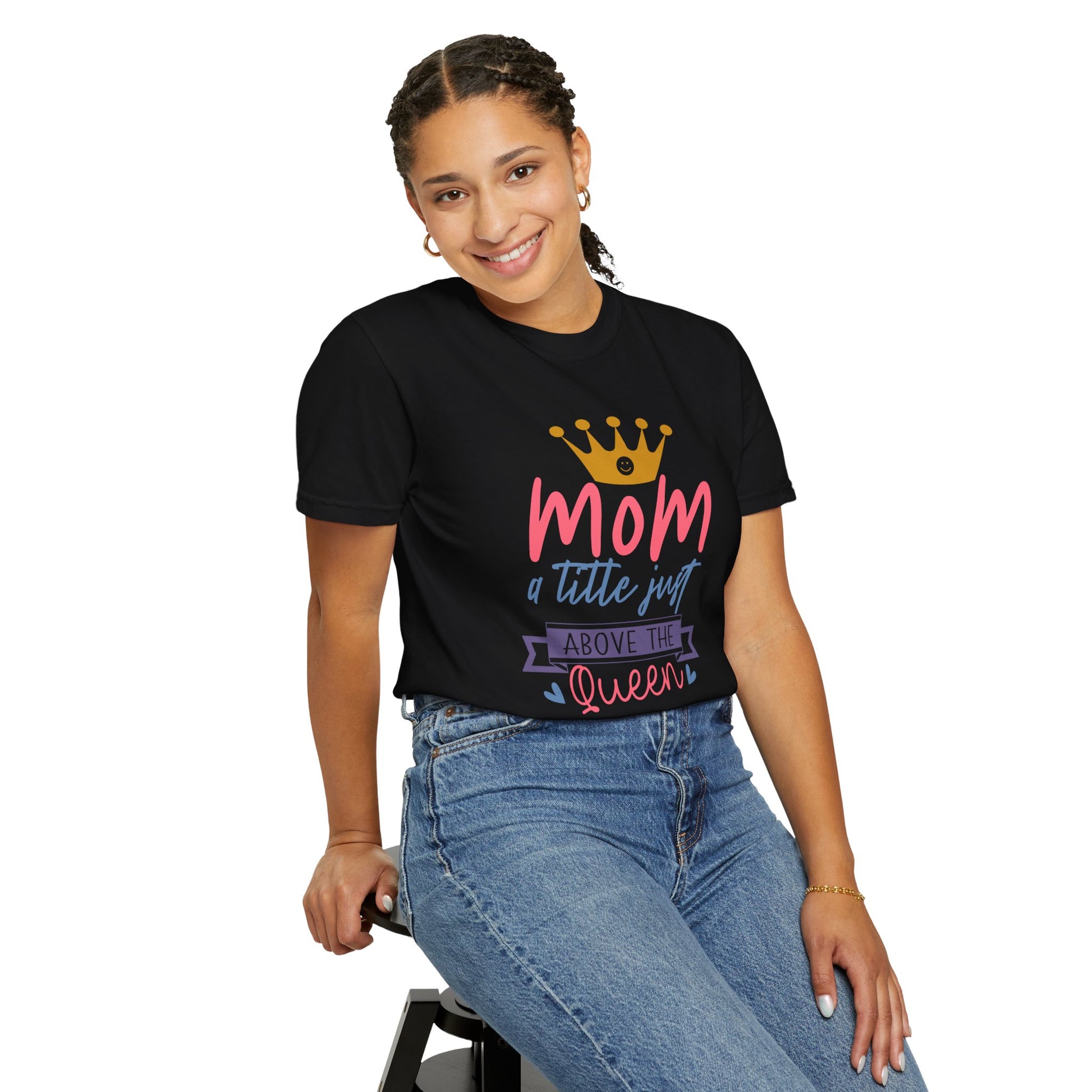 Clad in a Stylish Comfort with Unisex Comfort Colors 1717 Garment-Dyed T-Shirt featuring the phrase "mom just a little above queen," a person sits on a stool, smiling in their jeans. The ring-spun cotton brings an added layer of comfort to their joyful attire.