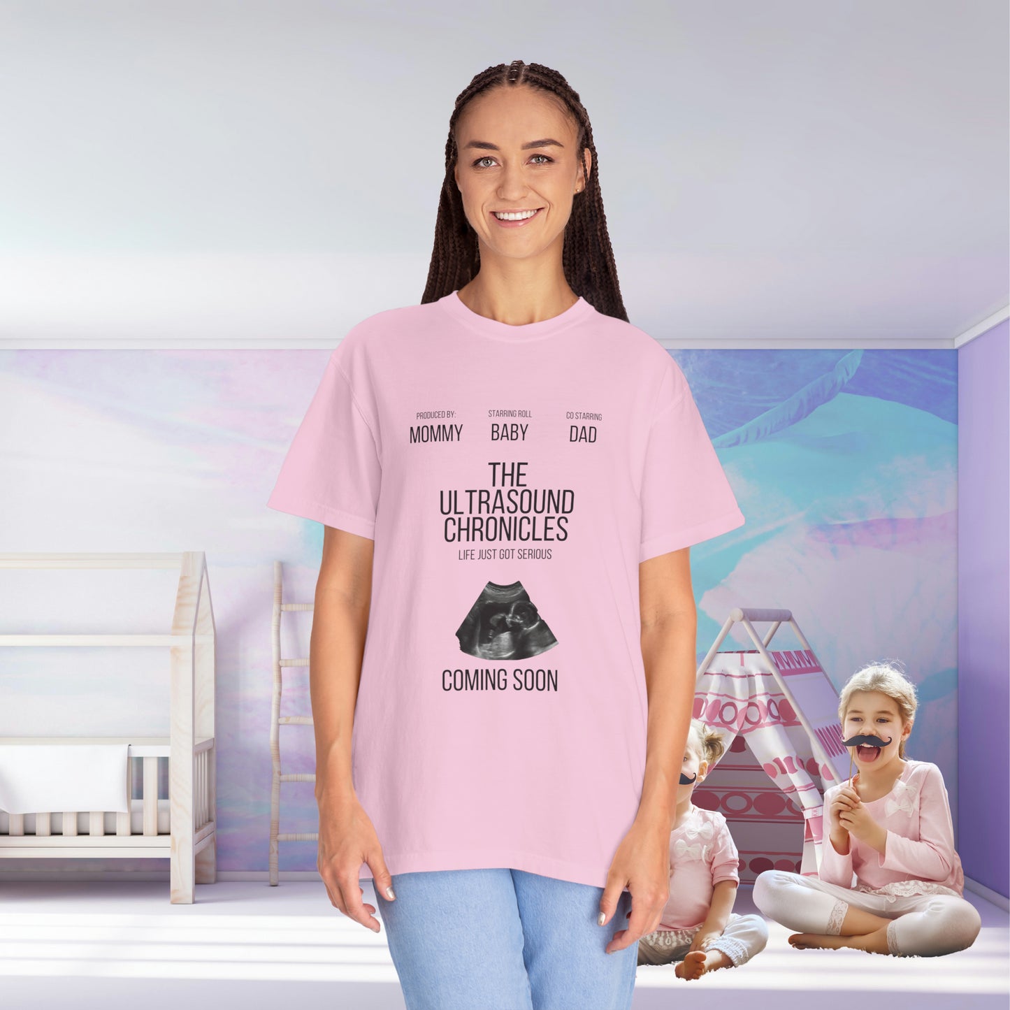 A woman in a pink Stylish Comfort with Unisex Comfort Colors 1717 Garment-Dye- t-shirt smiles in a nursery room; her shirt reads "the ultrasound chronicles: coming soon" with a hint for the upcoming gender reveal. Two playful children are in the