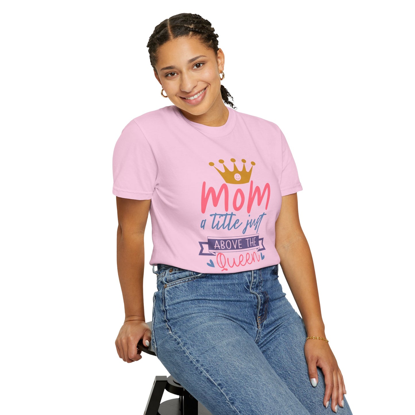 Wearing a pink Unisex Comfort Colors 1717 garment-dyed T-shirt crafted from comfortably soft ring-spun cotton, featuring the text "Mom just a little above queen" alongside a crown graphic, this person sits on a stool, smiling. It's like embracing royalty in one of those stylish Comfort Colors pieces.