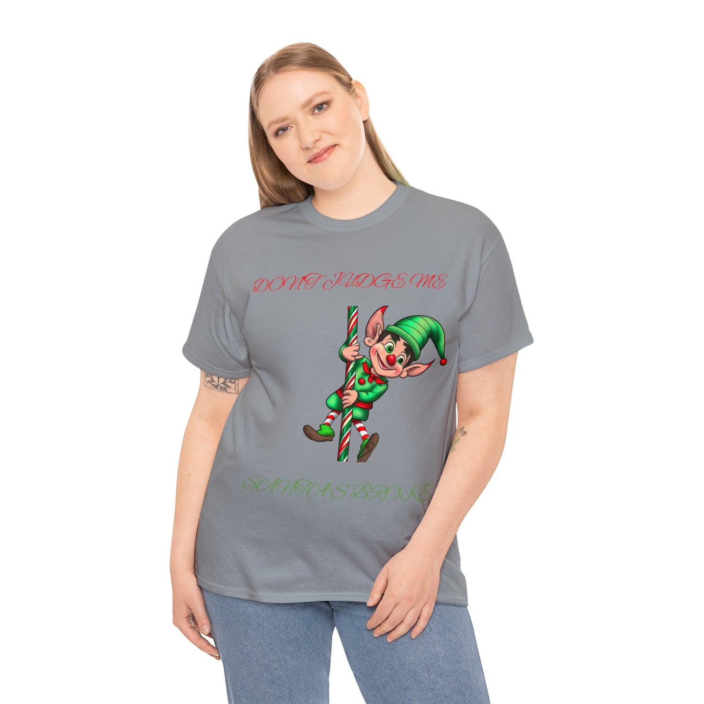 A person is wearing the Unisex Heavy Cotton- Don't Judge Me Elf T-shirt, featuring a classic fit in gray with a festive elf graphic and the text "DON'T JUDGE ME SANTA HASN'T," made from 100% cotton for a comfortable and sustainable option.