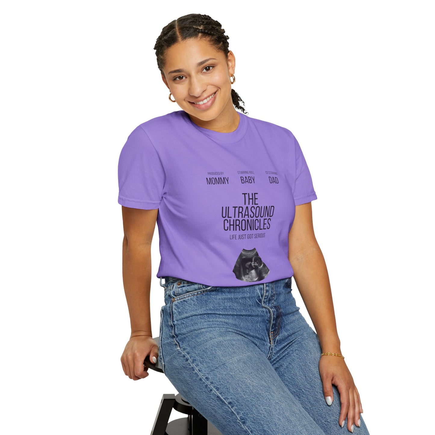 A person in a pair of jeans and a "Stylish Comfort with Unisex Comfort Colors 1717 Garment-Dye T-shirt - The Ultrasound Chronicles" sits on a stool, smiling.