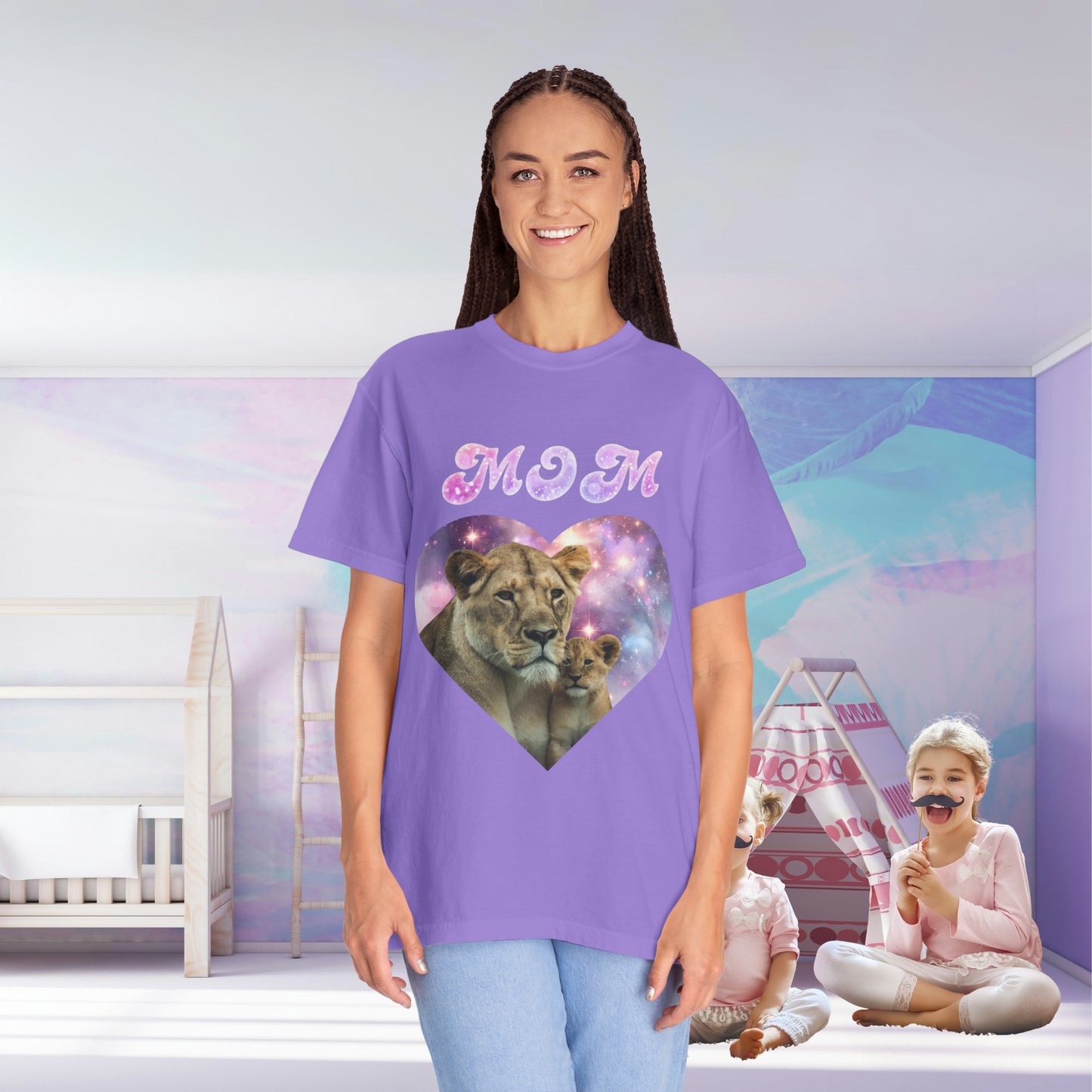 A mom in a Stylish Comfort with Unisex Comfort Colors 1717 Garment-Dye- T-shirt- Heart of the Cosmos featuring a graphic of a lioness and cub inside a cosmic heart, standing in a colorful room with her child playing in the background.