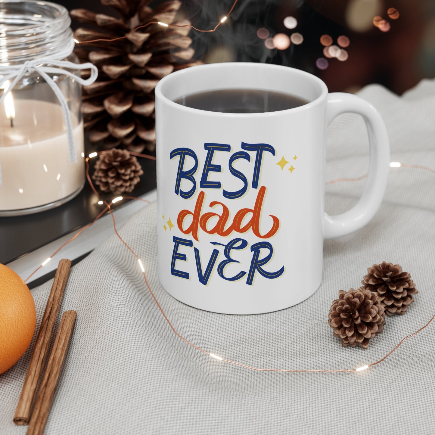 A white 11oz CeramicMug- best dad joke with "Best Dad Ever" written in colorful text sits on a fabric surface, surrounded by a candle, pinecones, cinnamon sticks, an orange, and decorative lights—the perfect Father's Day gift alongside a cozy t-shirt for the best father.