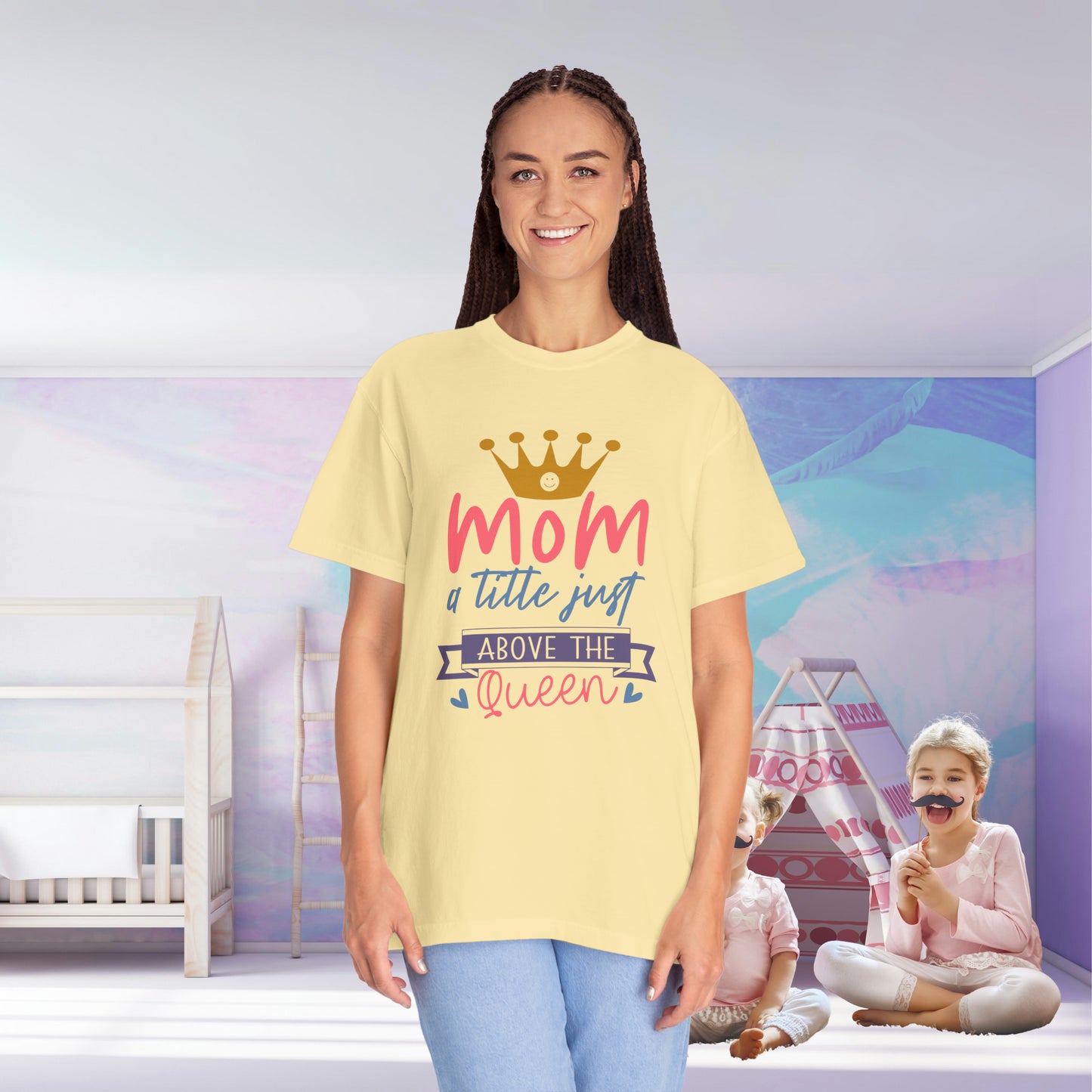 In a child's room, a woman stands wearing a Stylish Comfort with Unisex Comfort Colors 1717 Garment-Dyed T-Shirt featuring the text "mom just a little above queen." The garment-dyed, ring-spun cotton fabric radiates comfort while two children play joyfully in the background.