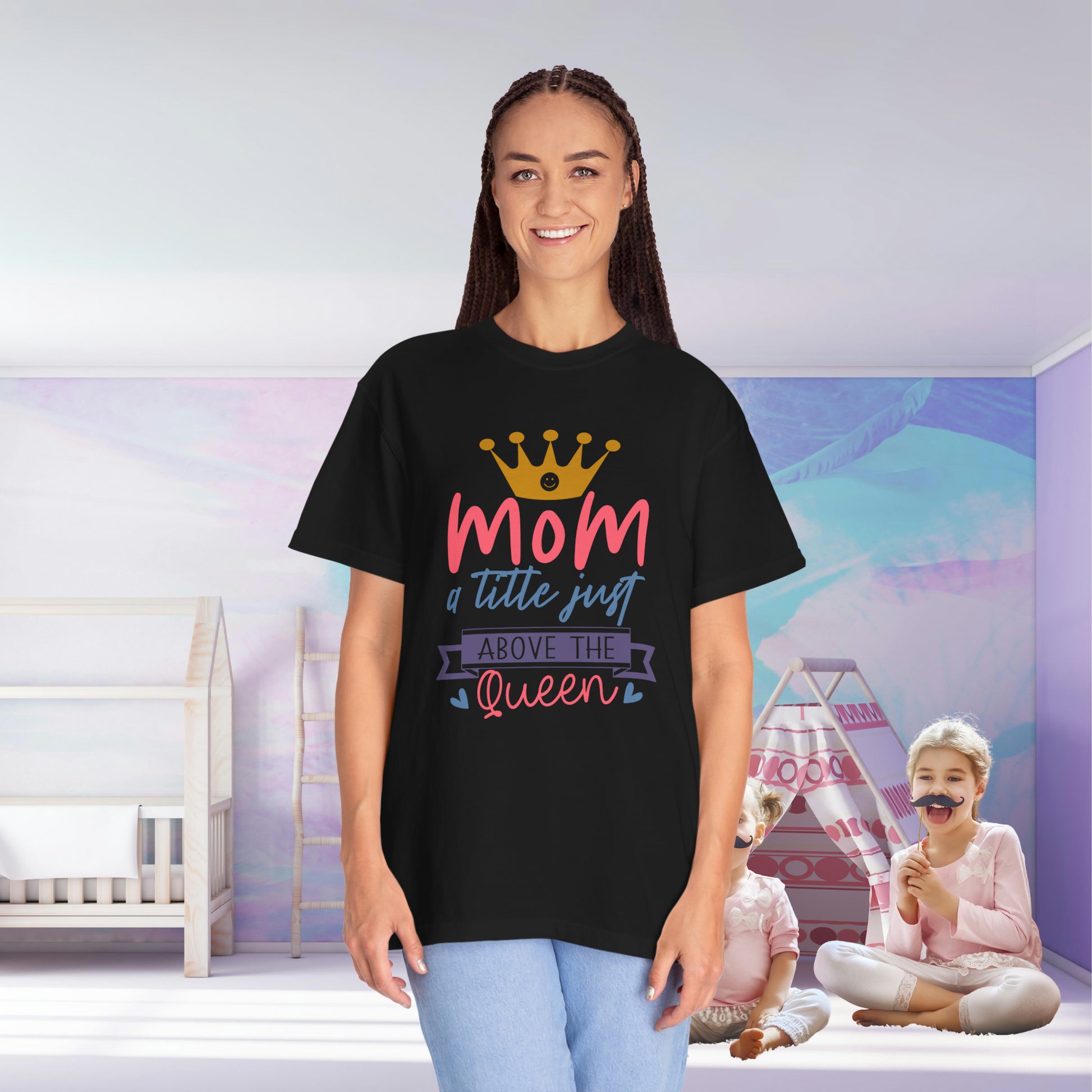 A woman wearing a Stylish Comfort with Unisex Comfort Colors 1717 Garment-Dyed T-Shirt, adorned with the text "mom just a little above queen," stands in a room while her two children play joyfully in the background.