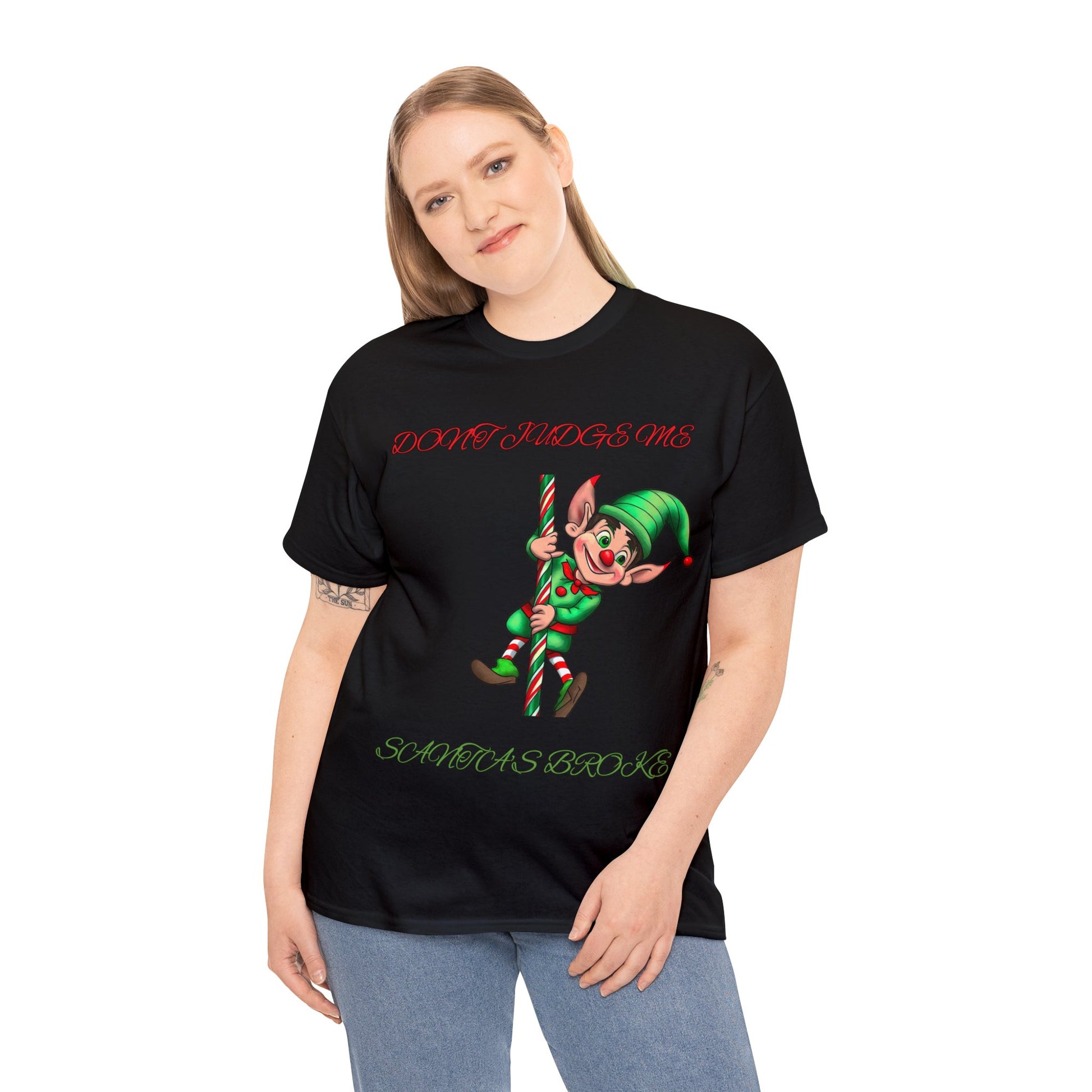 A person wears the Unisex Heavy Cotton- Don't Judge Me Elf T-shirt in a classic fit black design made from 100% cotton. This shirt showcases an elf graphic with the phrases "DON'T JUDGE ME" and "SANTA'S BROKEN," offering a sustainable fashion statement ideal for the holiday season.