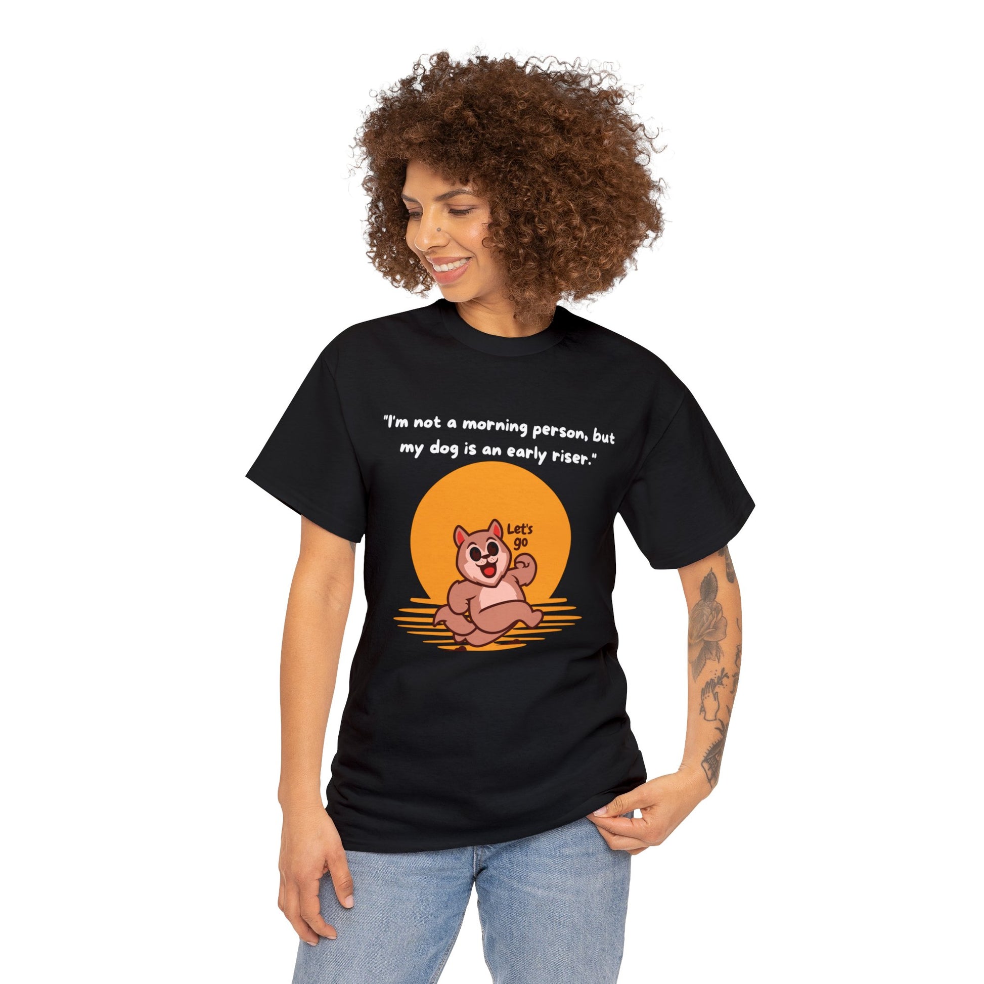 A person sports the Unisex Heavy Cotton Tee in a classic black fit, showcasing a cartoon dog and the message, "I'm not a morning person, but my dog is an early riser." Crafted from sustainable 100% cotton, this shirt blends comfort with humor for every dog lover.