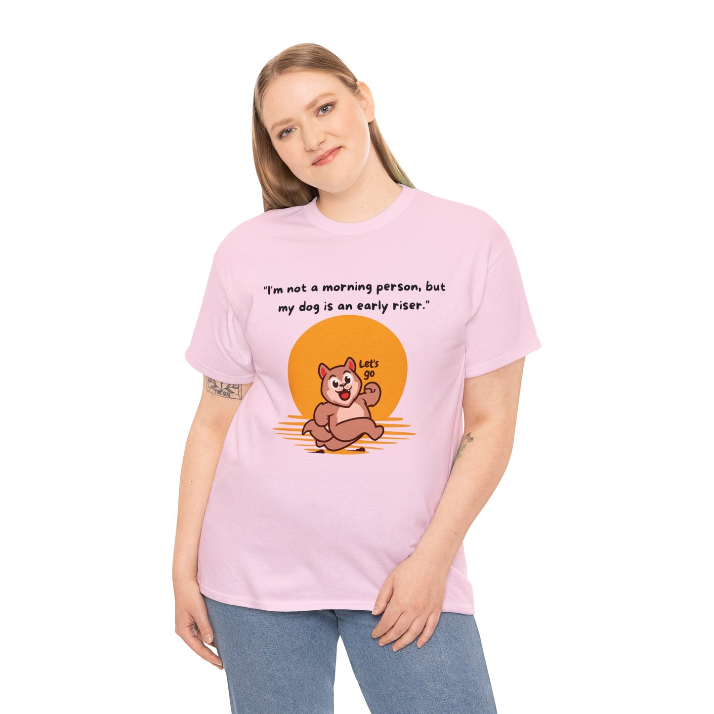 A person is wearing a Unisex Heavy Cotton Tee that showcases a cartoon dog and the phrase, "I'm not a morning person, but my dog is an early riser," in a classic fit pink design made from 100% cotton.