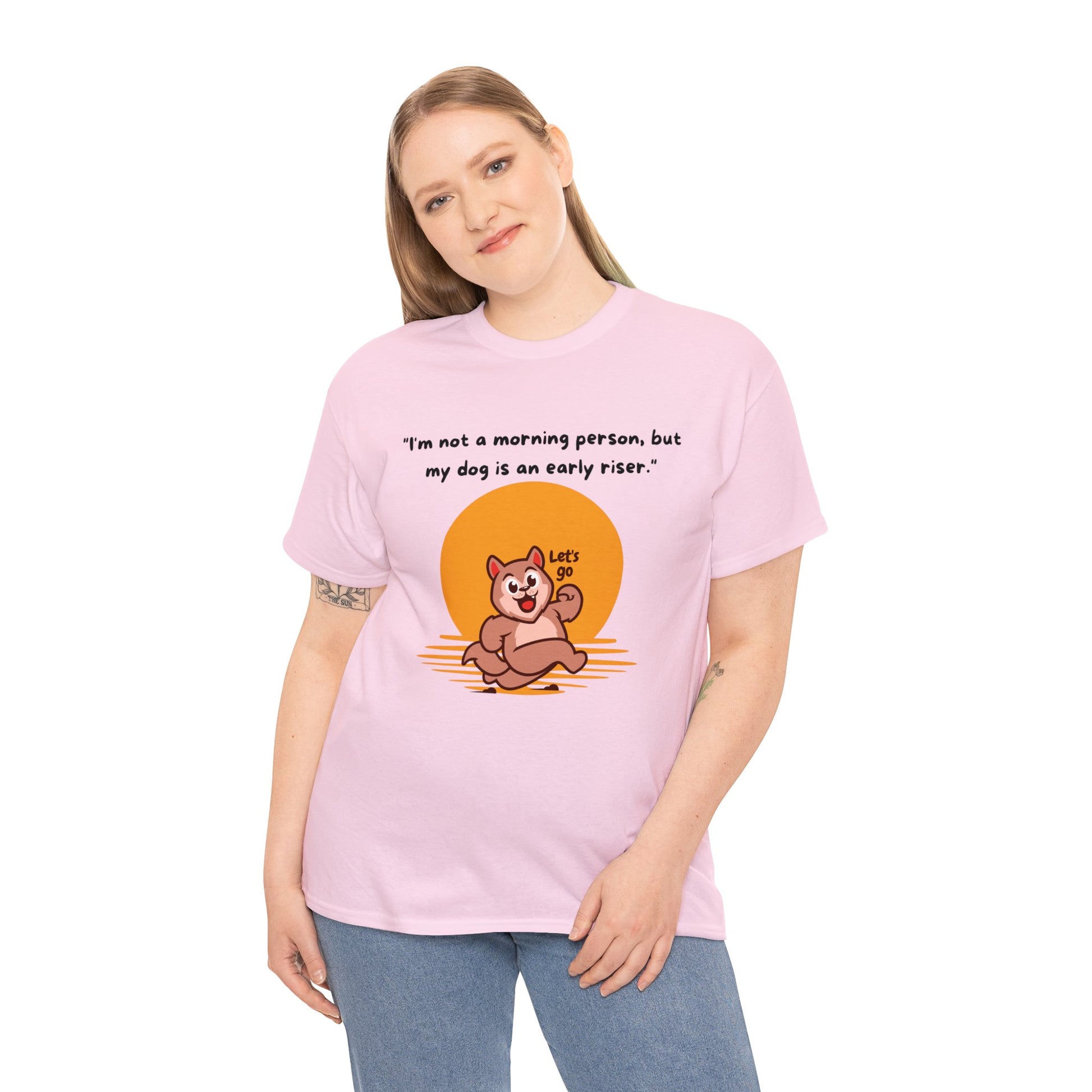 A person is wearing a Unisex Heavy Cotton Tee that showcases a cartoon dog and the phrase, "I'm not a morning person, but my dog is an early riser," in a classic fit pink design made from 100% cotton.