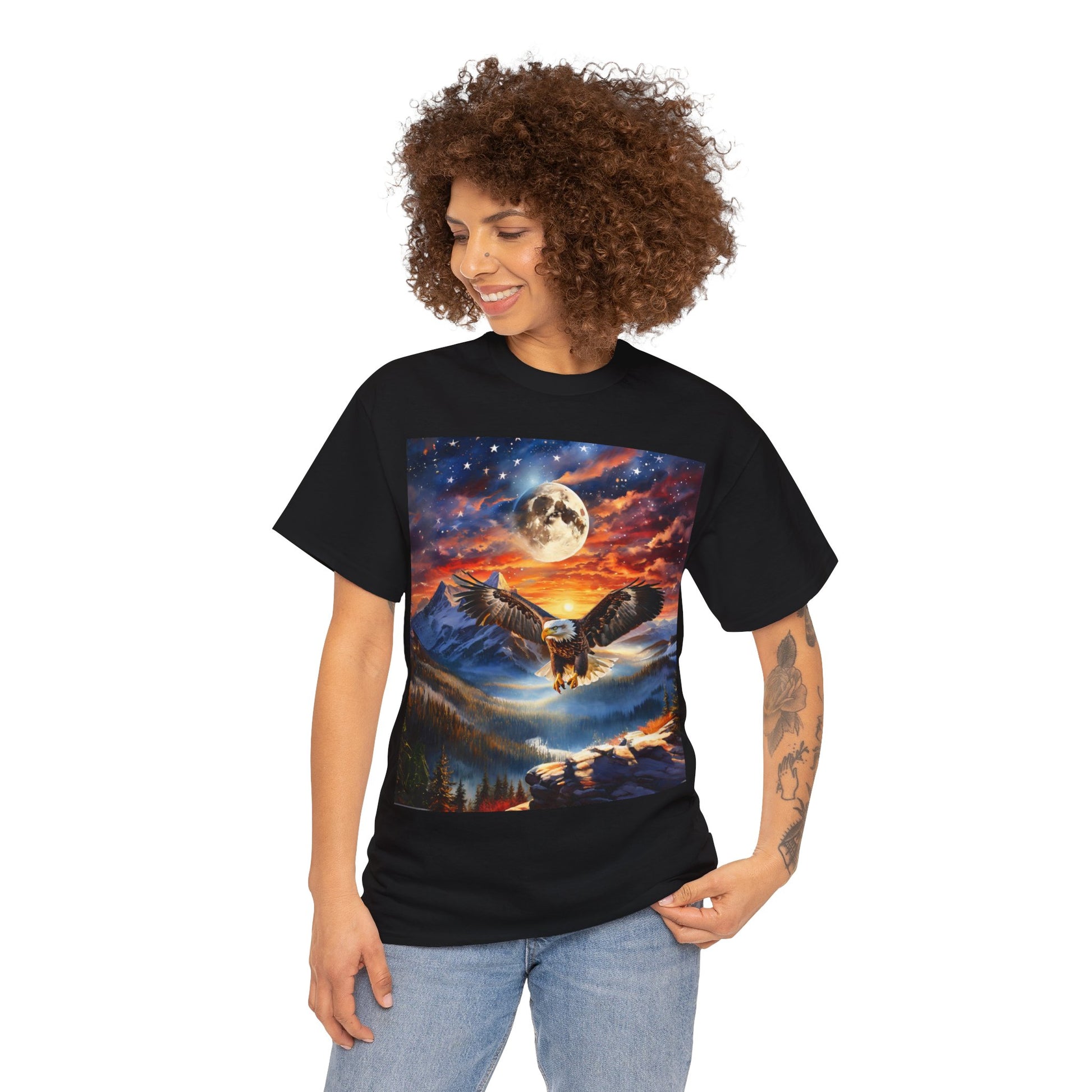 Person wearing a black Unisex Heavy Cotton Tee - Stars, Stripes, and Soaring Spirit: A Bald Eagle's Tribute with an owl in flight graphic against a moonlit mountain landscape, crafted from sustainably sourced cotton, standing and smiling.