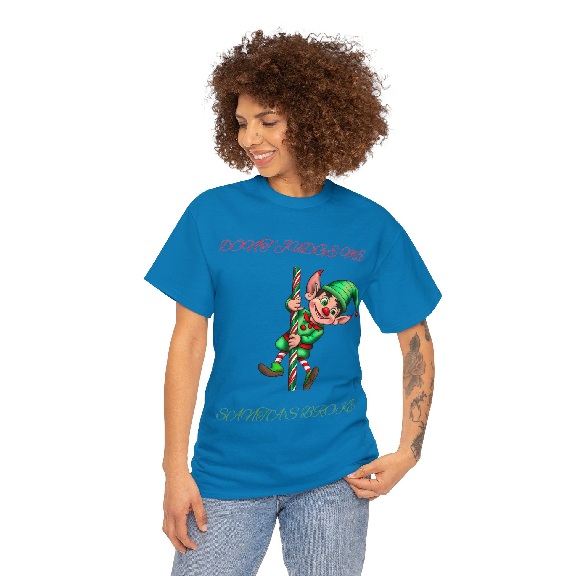 Wearing the Unisex Heavy Cotton- Don't Judge Me Elf T-shirt, a person sports a blue top adorned with a cartoon elf holding a candy cane and quirky text like "Don't judge me" and "Santa's Broke." Crafted from 100% cotton in a classic fit, this shirt blends sustainable fashion with lighthearted holiday fun.