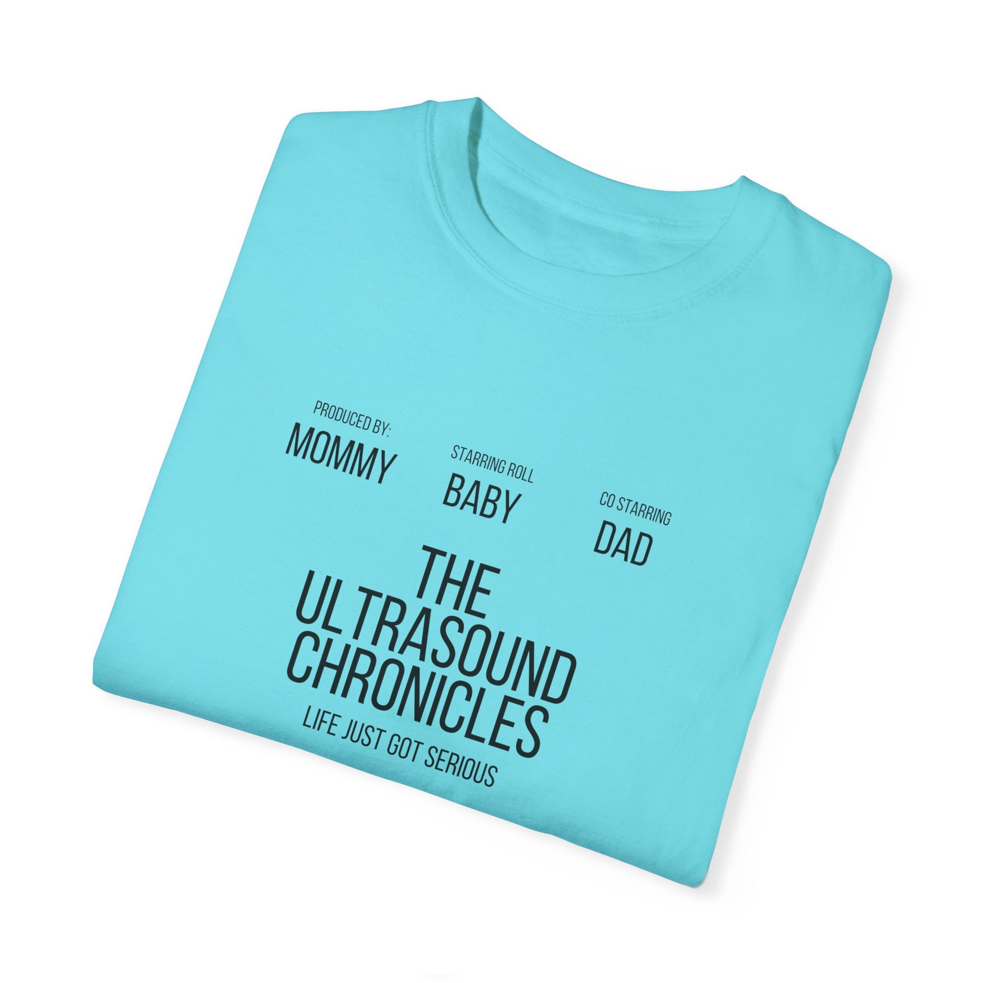 A folded Stylish Comfort with Unisex Comfort Colors 1717 Garment-Dye- T-shirt- The Ultrasound Chronicles in light blue with text "produced by mommy, starring baby, co-starring dad" printed in blue is perfect for a gender reveal.