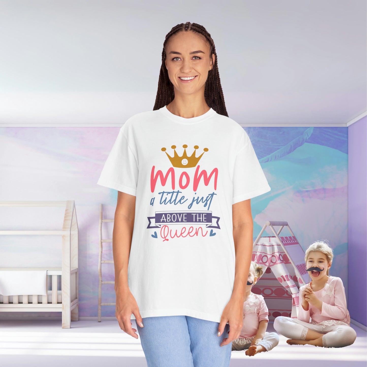 A woman stands in a room wearing a Stylish Comfort Unisex Comfort Colors 1717 Garment-Dyed T-Shirt that reads "Mom just a little above queen," crafted from soft ring-spun cotton. In the background, her two children play happily.