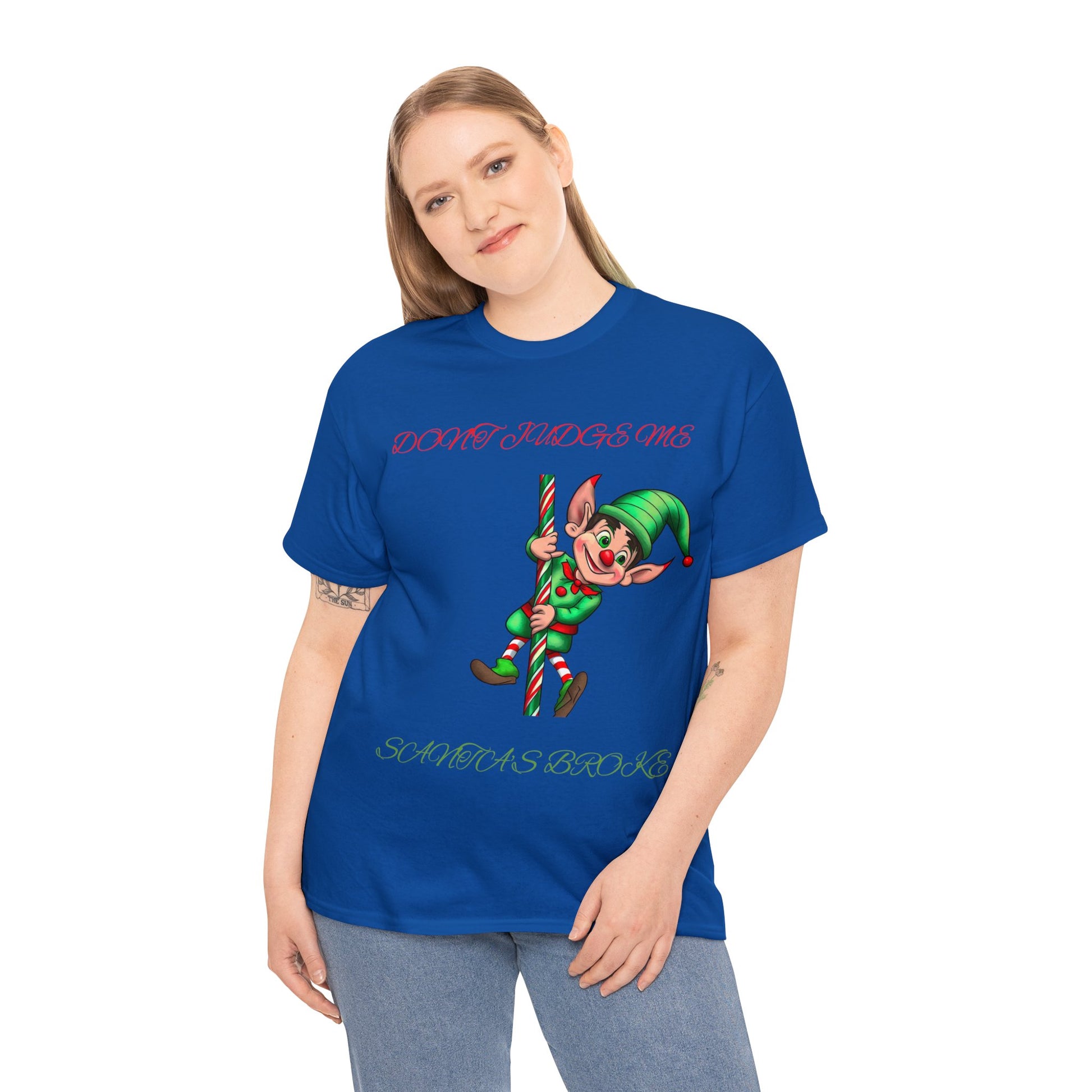 A person wearing an Unisex Heavy Cotton T-shirt with a festive elf design and the text "DON'T JUDGE ME SANTA'S BROKE" on the front.