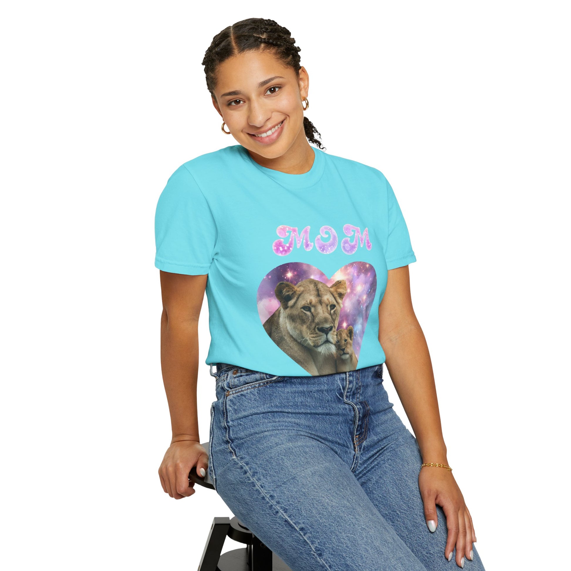 A person sits on a stool wearing the Stylish Comfort with Unisex Comfort Colors 1717 Garment-Dye T-shirt in turquoise, featuring a lioness and the word "MOM" in sparkly letters. They're smiling with braided hair, embracing the comfort of 100% ring-spun cotton.