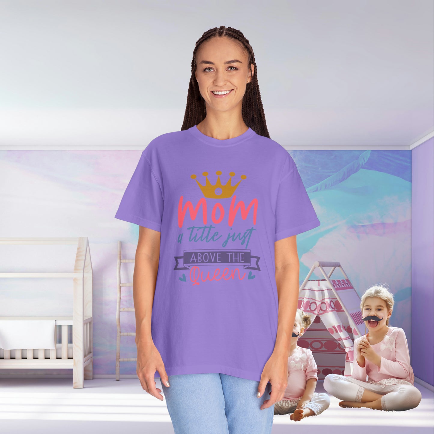 In a children's room, a woman wears a Stylish Comfort with Unisex Comfort Colors 1717 Garment-Dyed T-Shirt that reads "mom just a little above queen" in purple. Meanwhile, two children sit on the floor, one with face paint, enjoying the cozy feel of their ring-spun cotton outfits.