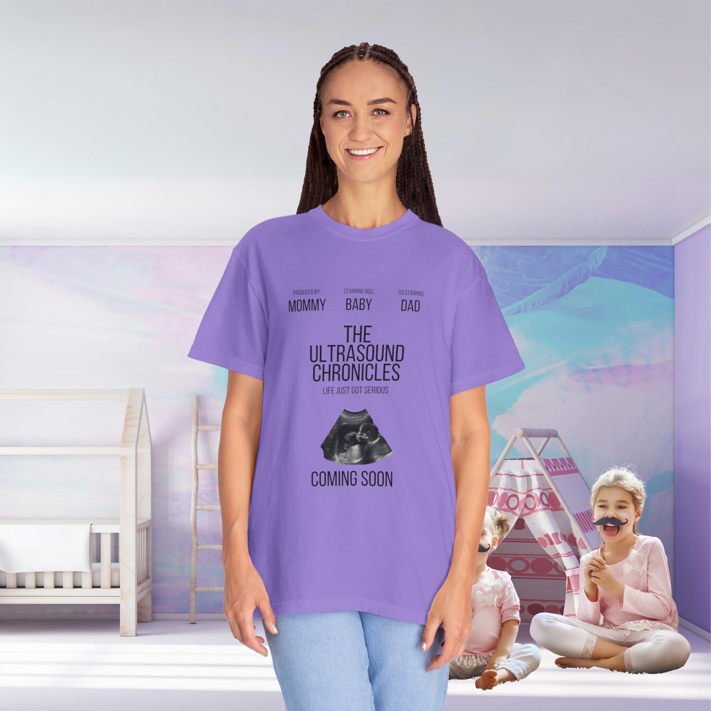 A new mom in a Stylish Comfort with Unisex Comfort Colors 1717 Garment-Dye- T-shirt- The Ultrasound Chronicles stands smiling in a nursery, with a child playing in the background. The shirt text announces her pregnancy with "The Ultrasound Chronicles: Coming Soon.