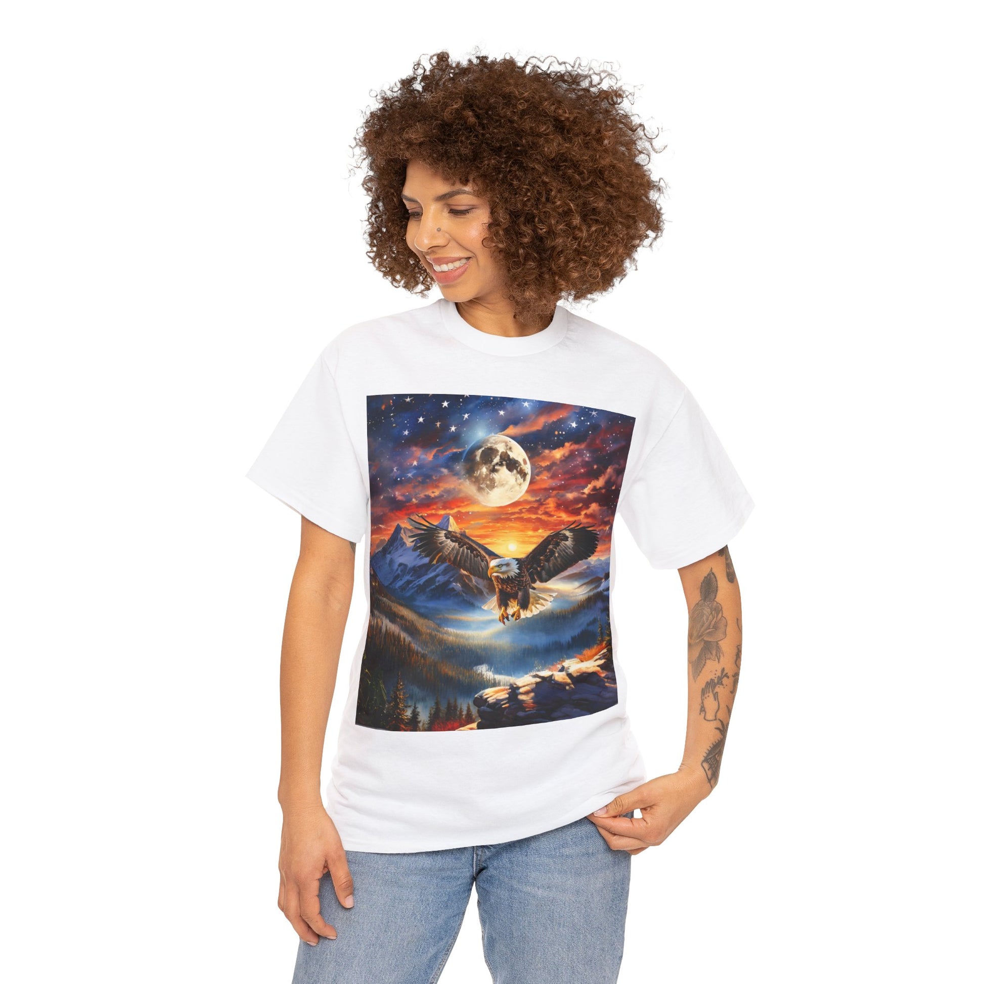 Person with curly hair wearing a Unisex Heavy Cotton Tee - Stars, Stripes, and Soaring Spirit: A Bald Eagle's Tribute featuring an illustrated scene of an owl flying over a mountainous landscape at night, made from sustainably sourced cotton.