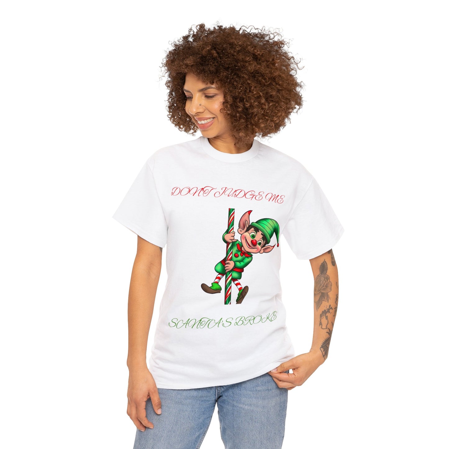 Showcasing both humor and comfort, a person wearing the Unisex Heavy Cotton- Don't Judge Me Elf T-shirt enjoys a classic fit. This white t-shirt features a cartoon elf holding a candy cane with the text "DON'T JUDGE ME SANTA'S BROKE," made from 100% cotton.