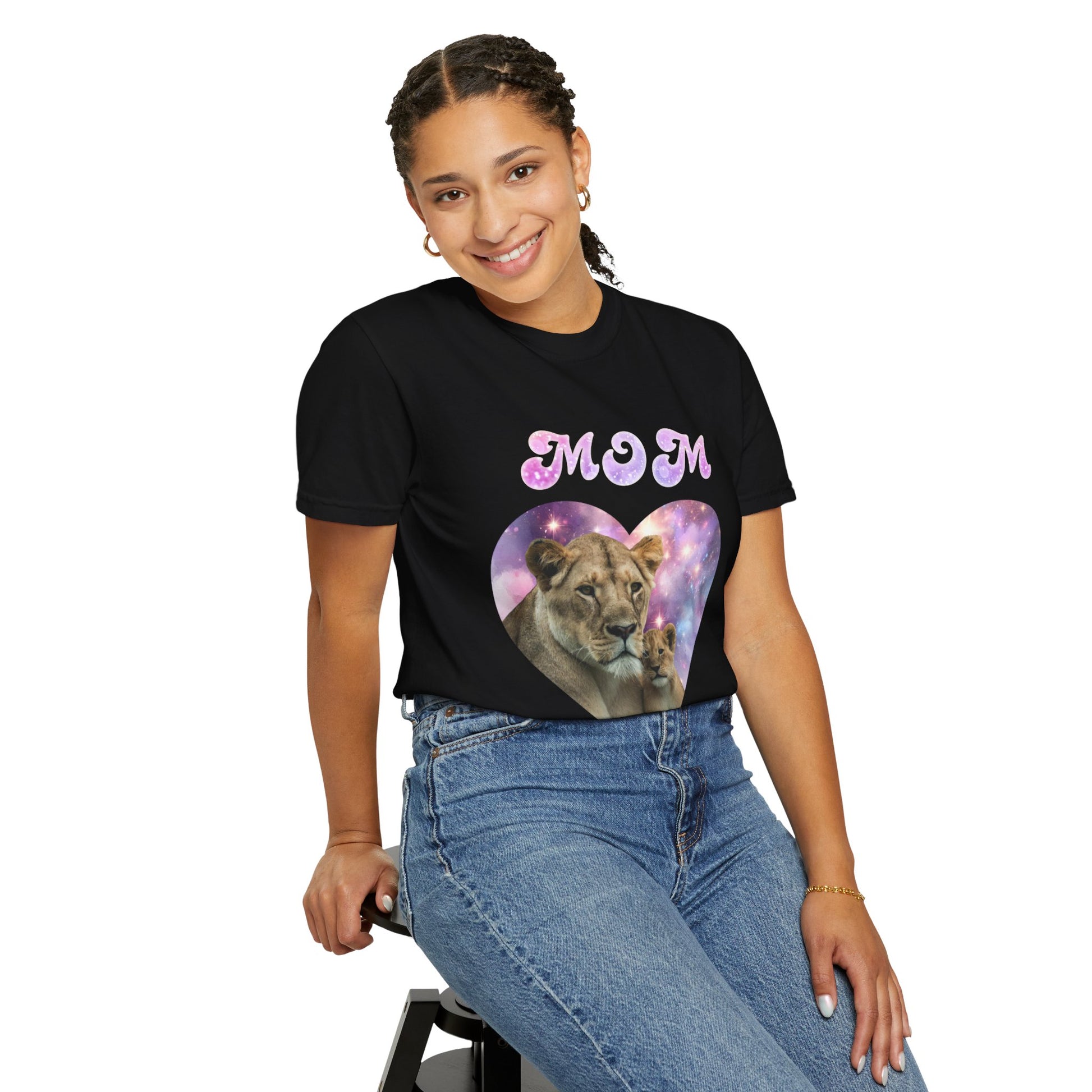 A young woman smiling, wearing a Stylish Comfort with Unisex Comfort Colors 1717 Garment-Dye- T-shirt- Heart of the Cosmos: Mom's Lioness Legacy, paired with blue jeans, sitting on a stool against a white background.