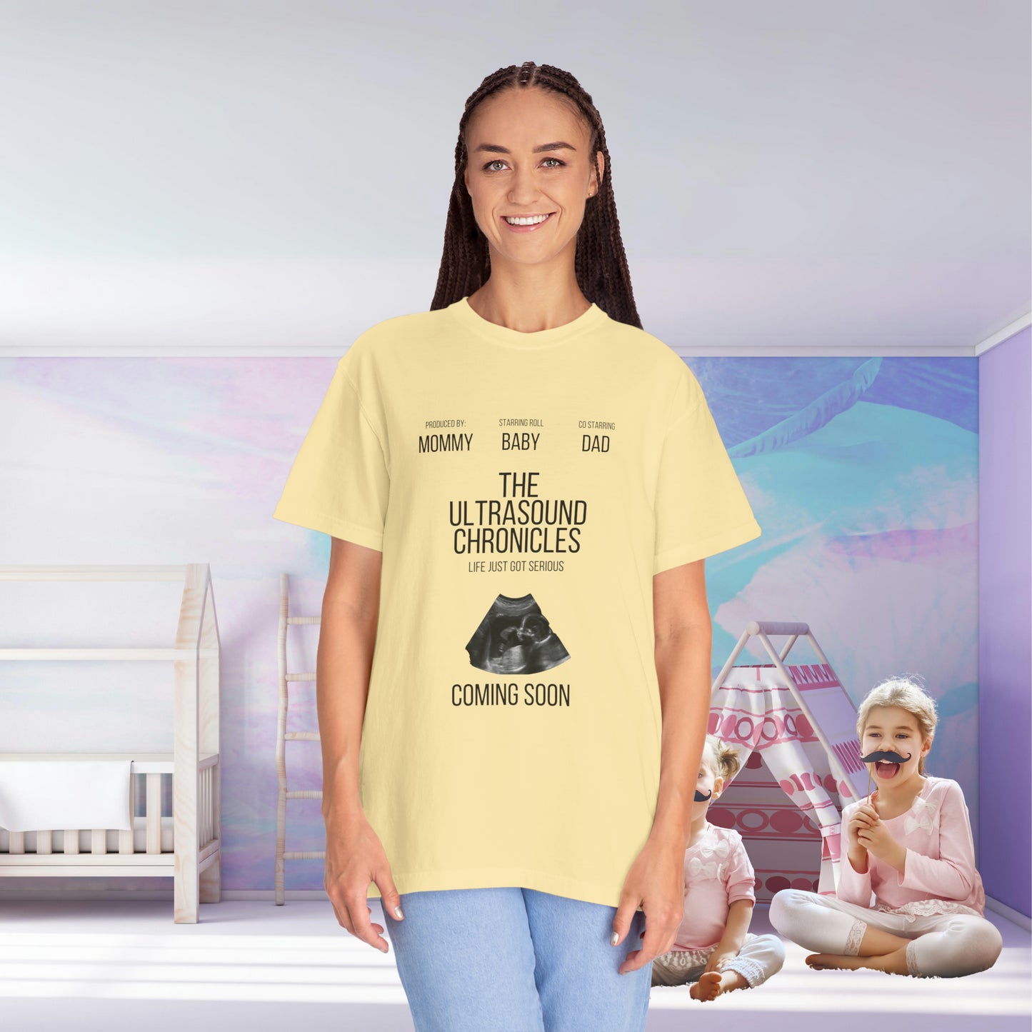 Woman in a yellow Stylish Comfort with Unisex Comfort Colors 1717 Garment-Dye T-shirt smiling at the camera, with text related to a baby shower on her shirt. In the background, a girl plays in a nursery room.