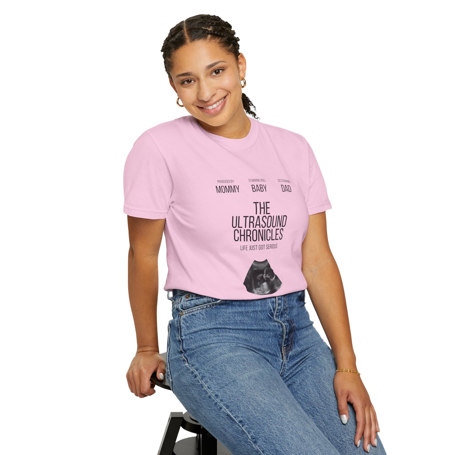 A woman sits on a stool against a white background, smiling as she wears the "Stylish Comfort with Unisex Comfort Colors 1717 Garment-Dye T-shirt - The Ultrasound Chronicles." The pink tee highlights an ultrasound image and embodies joy and anticipation with its soft ring-spun cotton fabric.