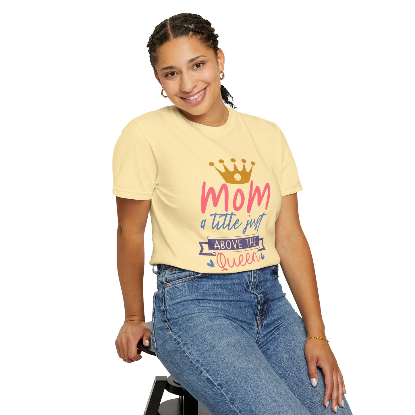 A person wearing a beige Unisex Comfort Colors 1717 Garment-Dyed T-Shirt from the "Stylish Comfort" collection, featuring the text "mom just a little above queen" and a crown graphic, is sitting on a stool and smiling.