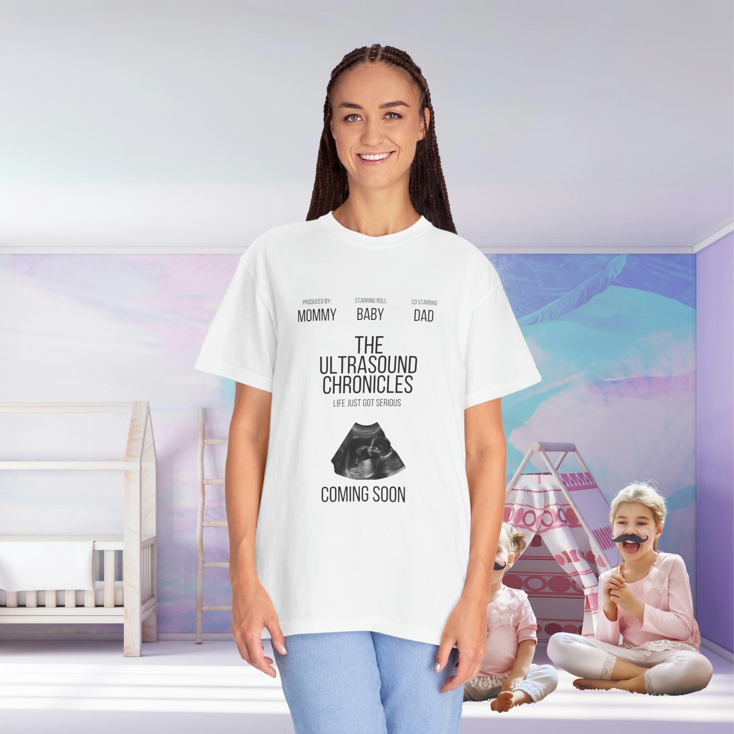 A smiling woman in a white Stylish Comfort with Unisex Comfort Colors 1717 Garment-Dye- T-shirt that reads "the ultrasound chronicles" with background images of a nursery ready for the newborn baby.