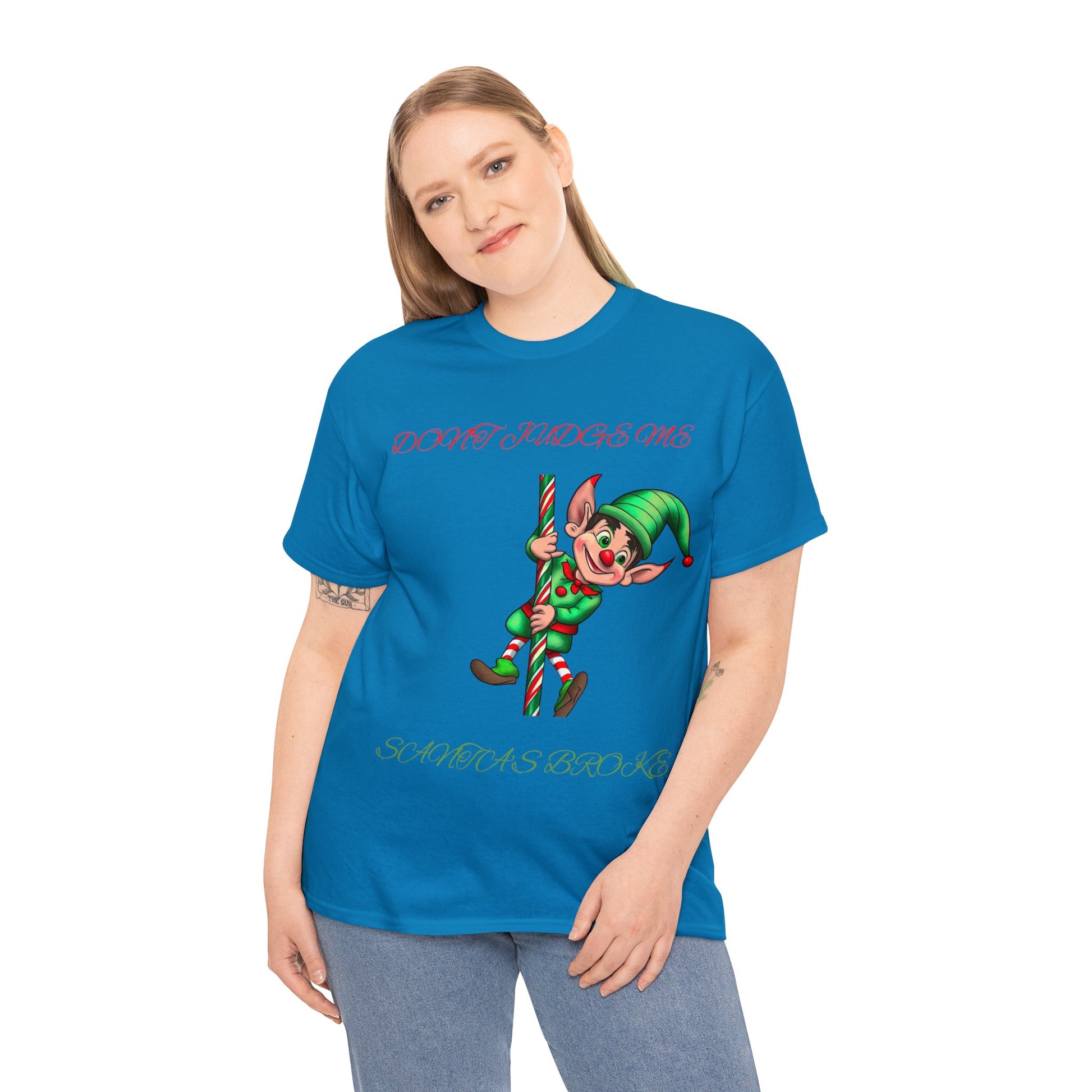 A person is wearing the Unisex Heavy Cotton- Don't Judge Me Elf t-shirt, which is sustainably made with a classic fit and 100% cotton, showcasing a cartoon elf and the words "DON'T JUDGE ME SANTA'S BROKEN.