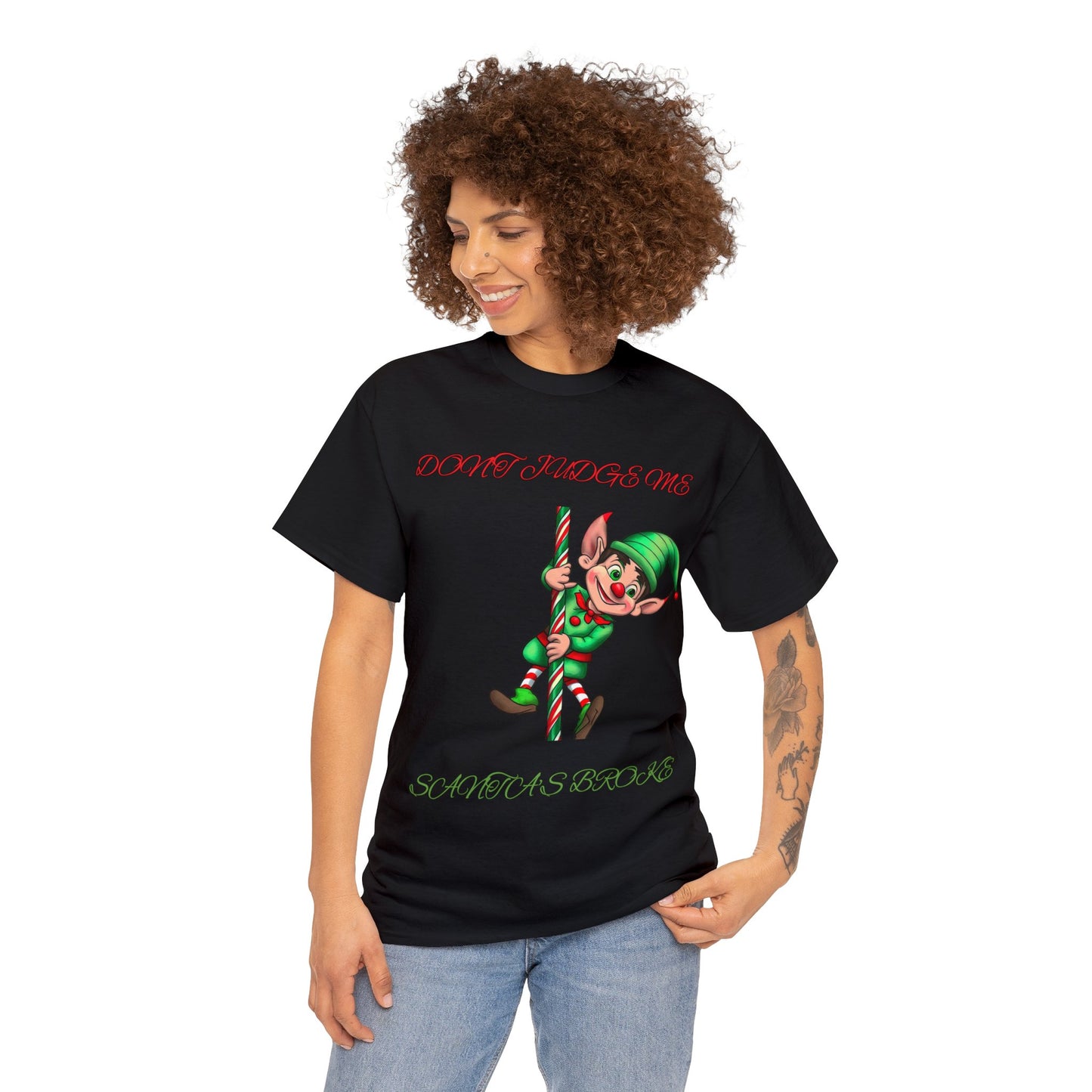 A person wearing the Unisex Heavy Cotton- Don't Judge Me Elf t-shirt in classic black, featuring 100% cotton fabric and a cartoon elf holding a candy cane accompanied by the text "Don't Judge Me Santa's Broke.