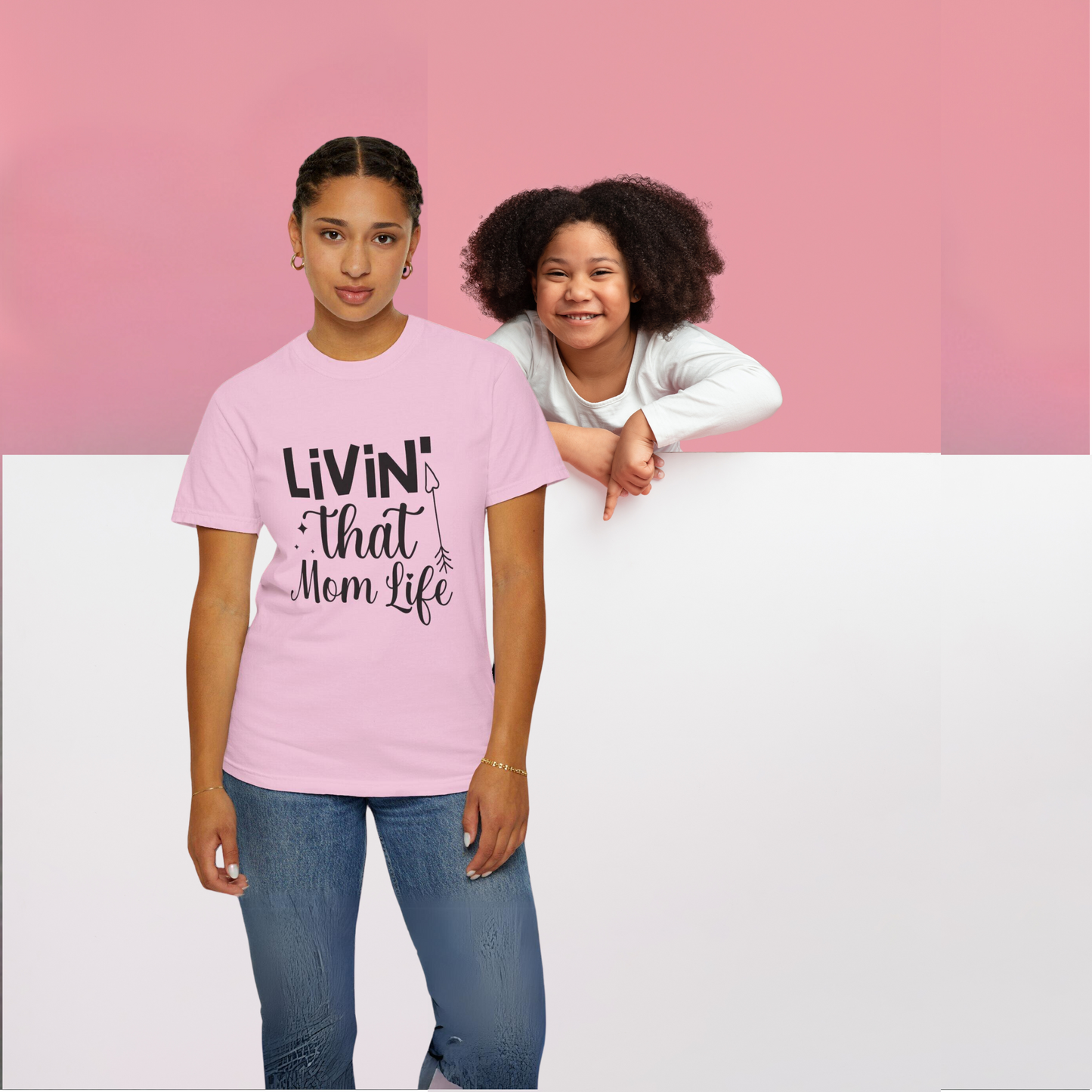 A woman wearing a pink "Stylish Comfort with Unisex Comfort Colors 1717 Garment-Dyed T-Shirt - Living that Mom Life" stands alongside a girl, both leaning against a white wall set against a two-tone pink and white backdrop.