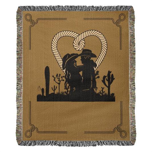 This 50x60 Heirloom Woven Blanket, made from 100% cotton yarn, showcases a distinct design featuring a silhouette of a cowboy-hat-wearing couple embracing in front of a heart formed by rope. The scene is enhanced with charming cacti and western motifs along the border.