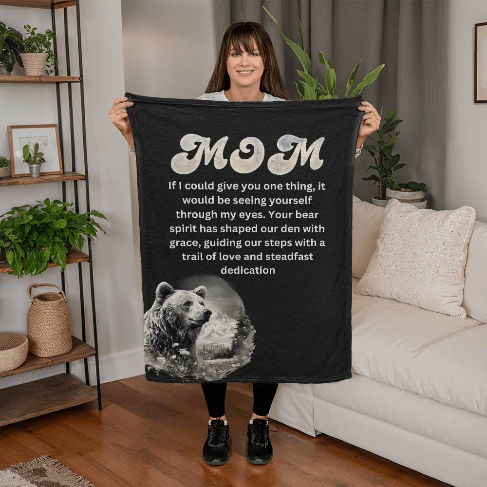 A woman smiles, holding up a Coral Fleece Blanket- Guardian of Love: Embracing Mom's Bear Spirit with "Mom" and a heartfelt Mothers Day message alongside a bear graphic, in a cozy living room setting.