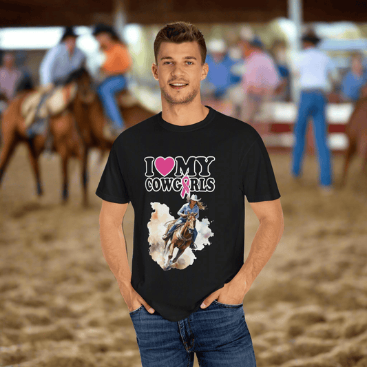 A man wearing a black "I Love My Cowgirls" T-shirt, designed for breast cancer awareness, stands in a sandy arena surrounded by people and horses. The stylish Comfort Colors 1717 unisex garment-dyed T-shirt, crafted from soft ring-spun cotton, perfectly complements his casual style amidst the lively scene.